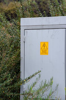 A gray electrical box surrounded by dense bushes with a warning sticker displayed prominently.